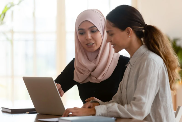 Two professional women sit at a desk, collaborating on a laptop. One woman, wearing a light pink hijab and a beige blazer, gestures towards the screen while speaking. The other woman, dressed in a white blouse, listens attentively with a slight smile. Natural light streams in from a large window in the background, creating a bright and welcoming workspace.